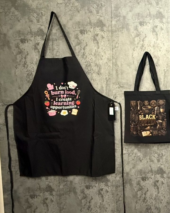 A black apron with a colorful kitchen print next to a printed black tote